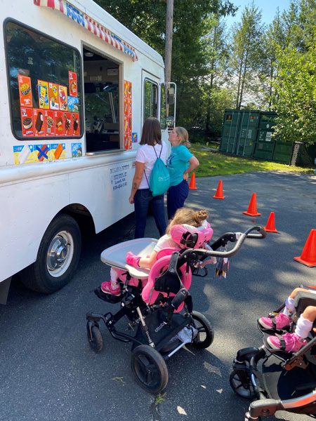 An ice cream truck with two ladies buying ice cream and kids sitting